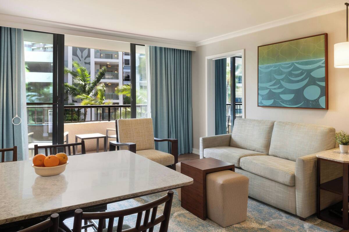 A hotel room with a dining table, chairs, a sofa, and a large window with curtains.