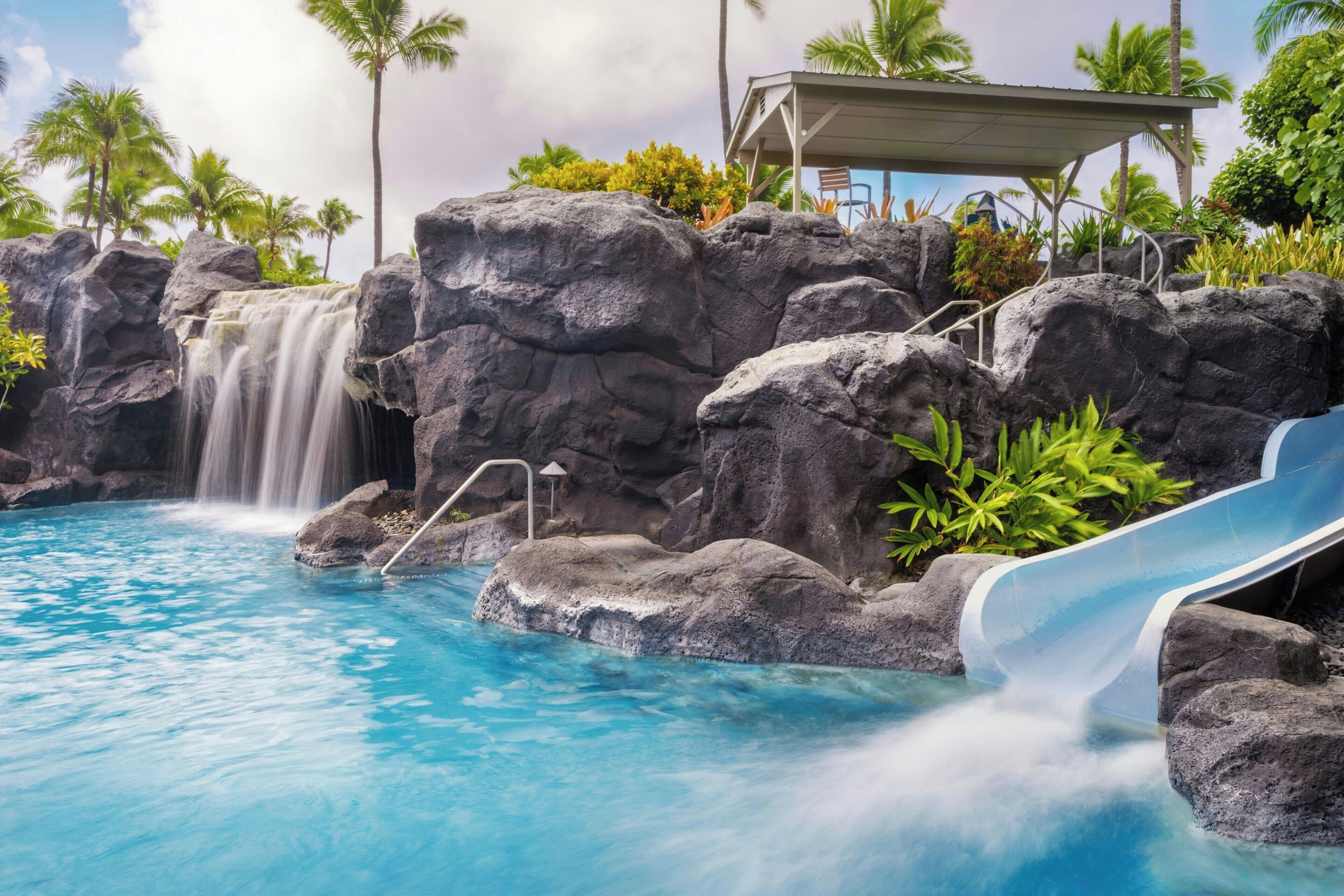 A pool with a waterfall, a slide, and a covered seating area with chairs.