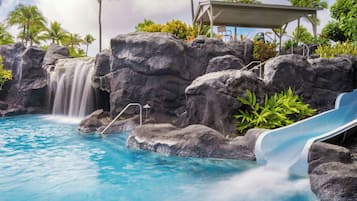 A pool with a waterfall, a slide, and a covered seating area with chairs.