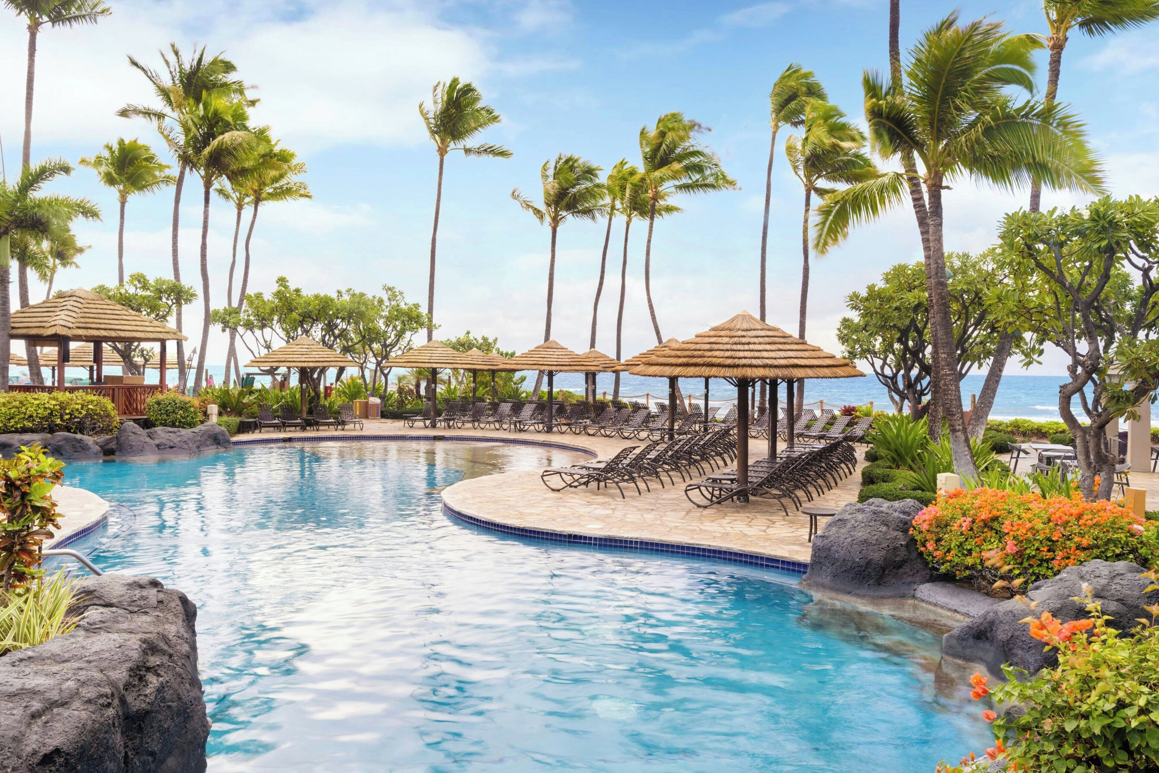 A pool area with lounge chairs, palm trees, and a view of the ocean.