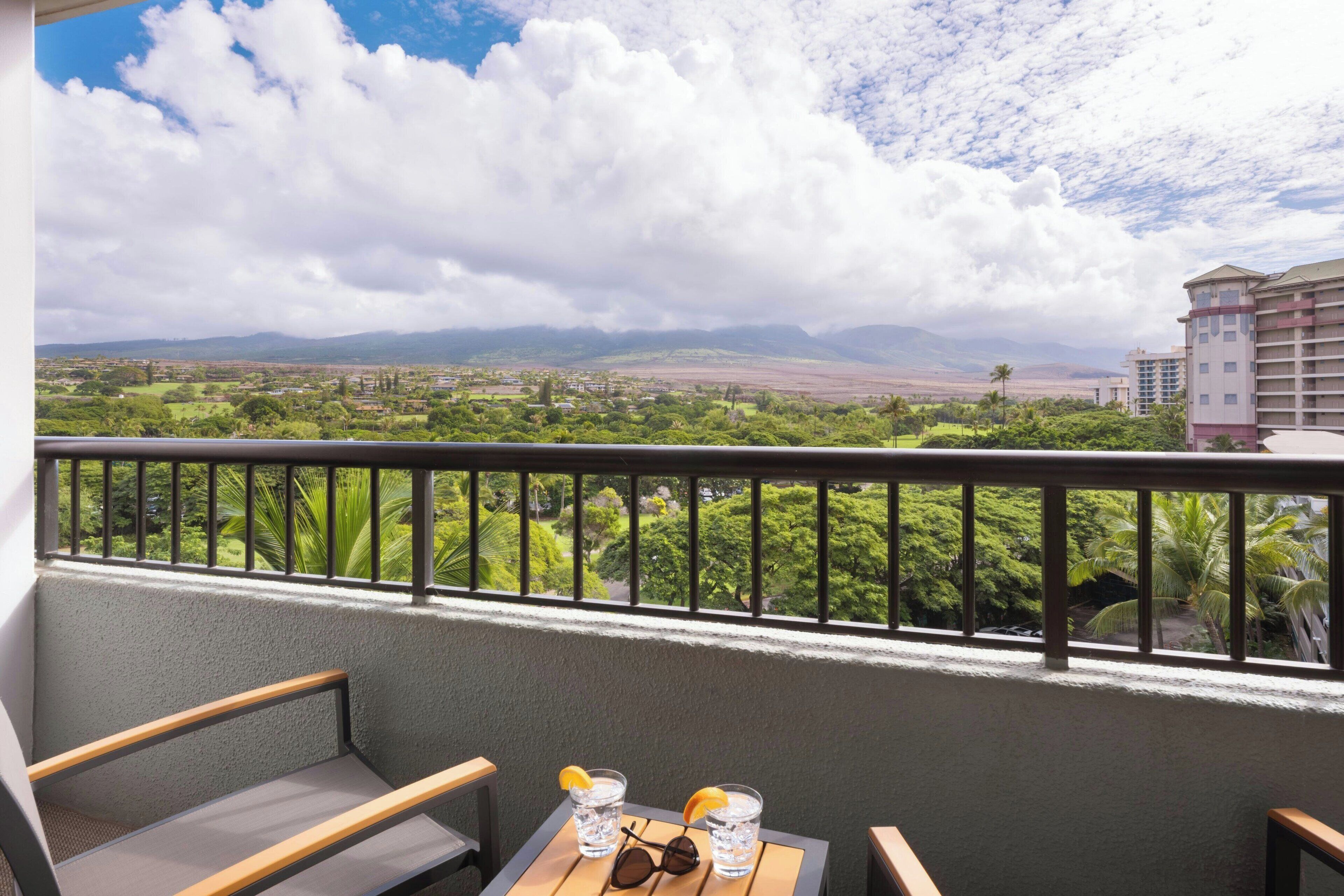 A balcony with a view of a cityscape and mountains, a table with glasses and sunglasses, and a bench.