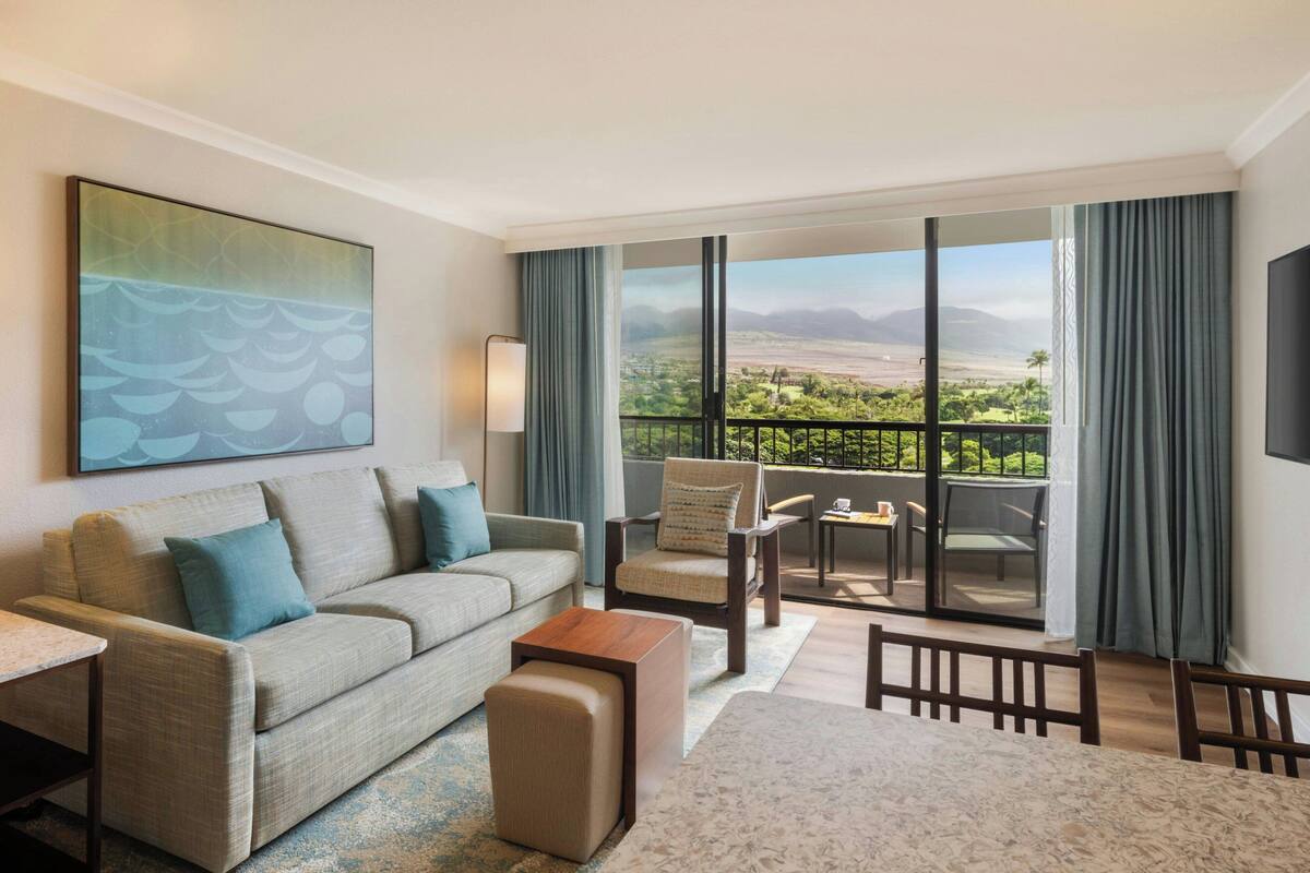 Een hotelkamer met balkon, een bank, een stoel, een tafel en een schilderij aan de muur.