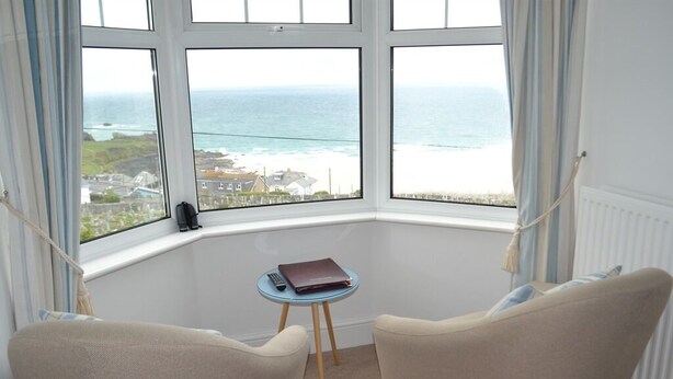 Una camera d'angolo con due poltrone beige, un tavolino rotondo con un telecomando e un libro, e vista sul mare e sugli edifici attraverso ampie finestre.