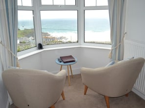 A corner room with two beige armchairs, a small round table with a remote control and a book, and a view of the sea and buildings through large windows. - Tregony Guest House (St Ives)