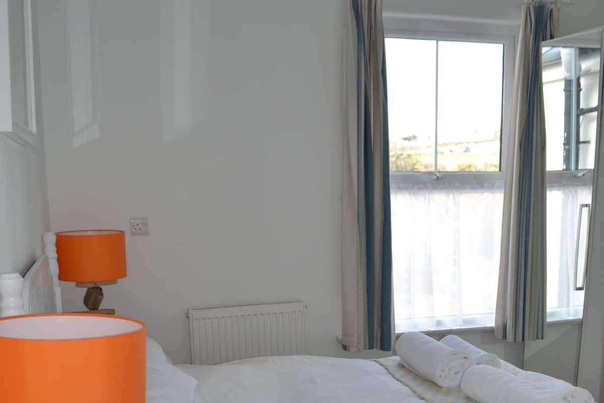 A bedroom with a bed, a nightstand with a lamp, a window with curtains, and a view of the outdoors.
