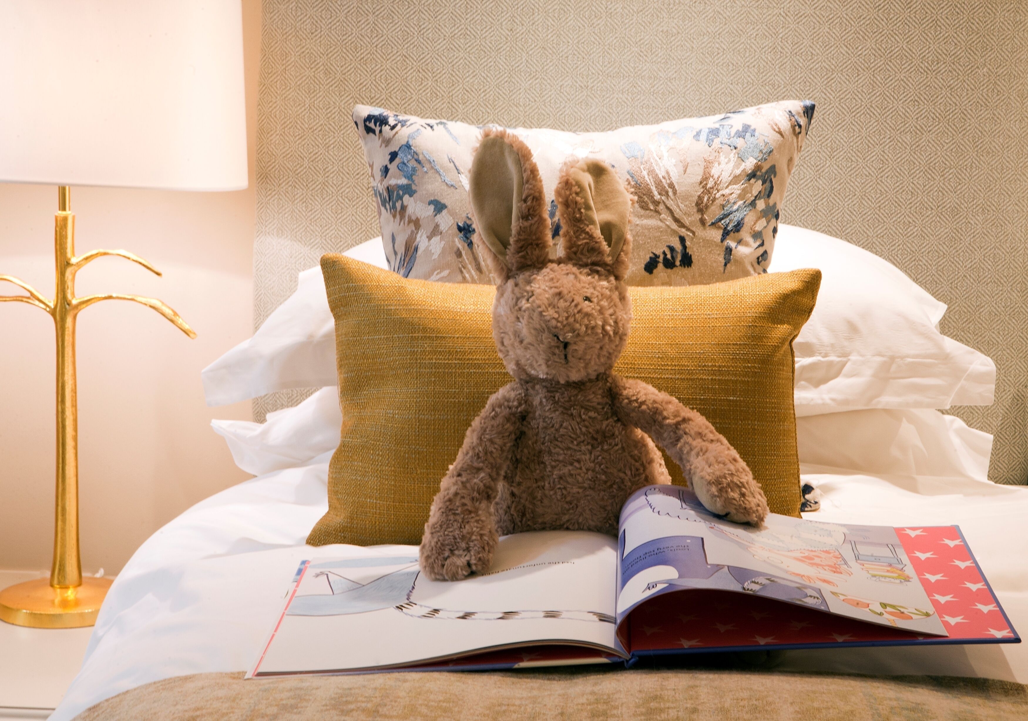 A plush toy on a bed with a book and a lamp.