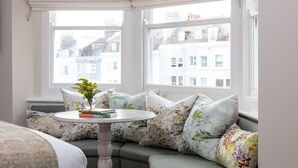 Una habitación con ventanal triangular, una mesa redonda con libros y un florero, y un sofá con cojines estampados.