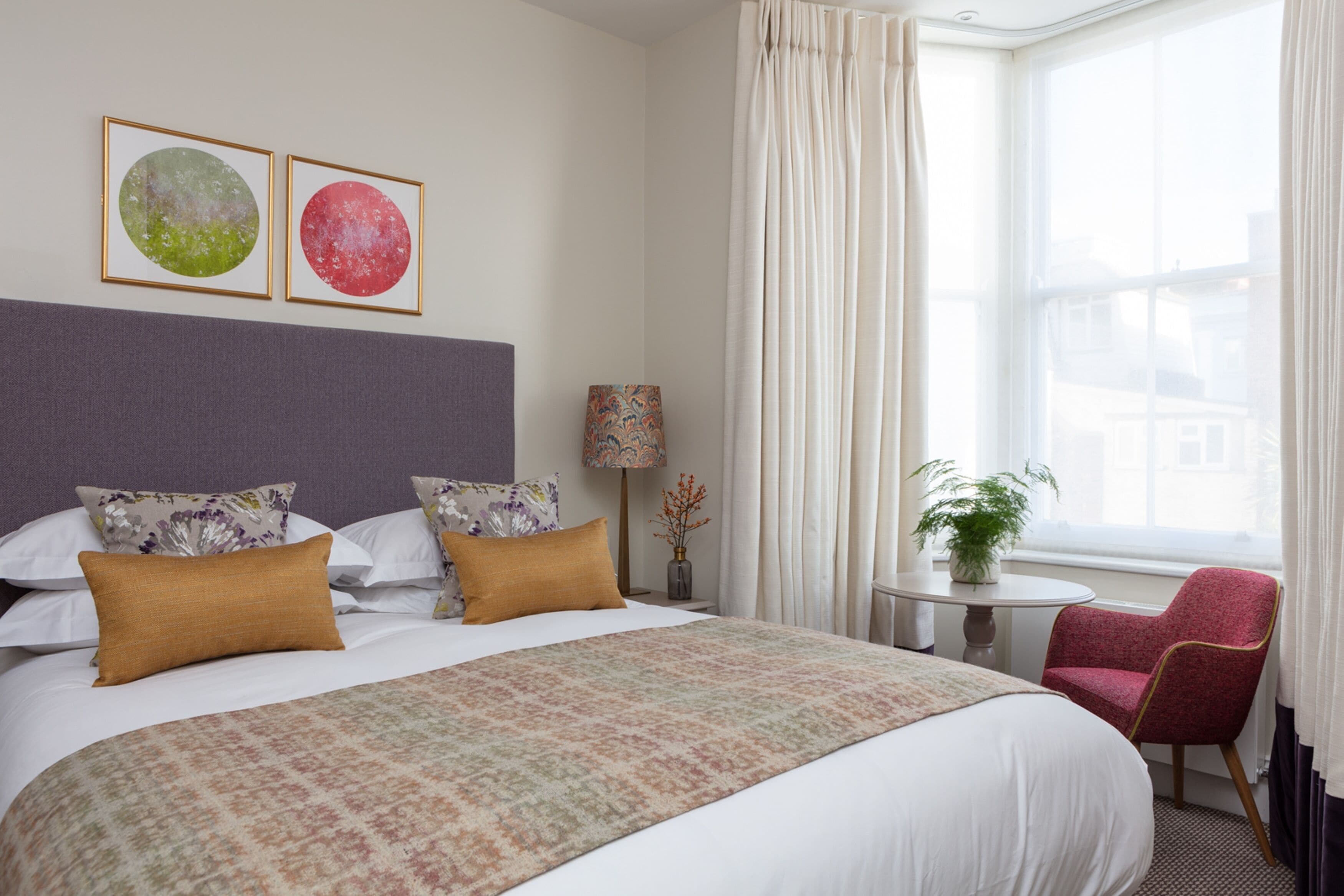 A hotel room with a large bed, two framed pictures on the wall, a small table with a lamp, a chair, and a window with curtains.