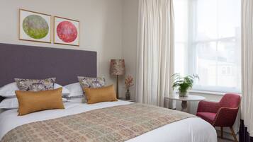 A hotel room with a large bed, two framed pictures on the wall, a small table with a lamp, a chair, and a window with curtains.