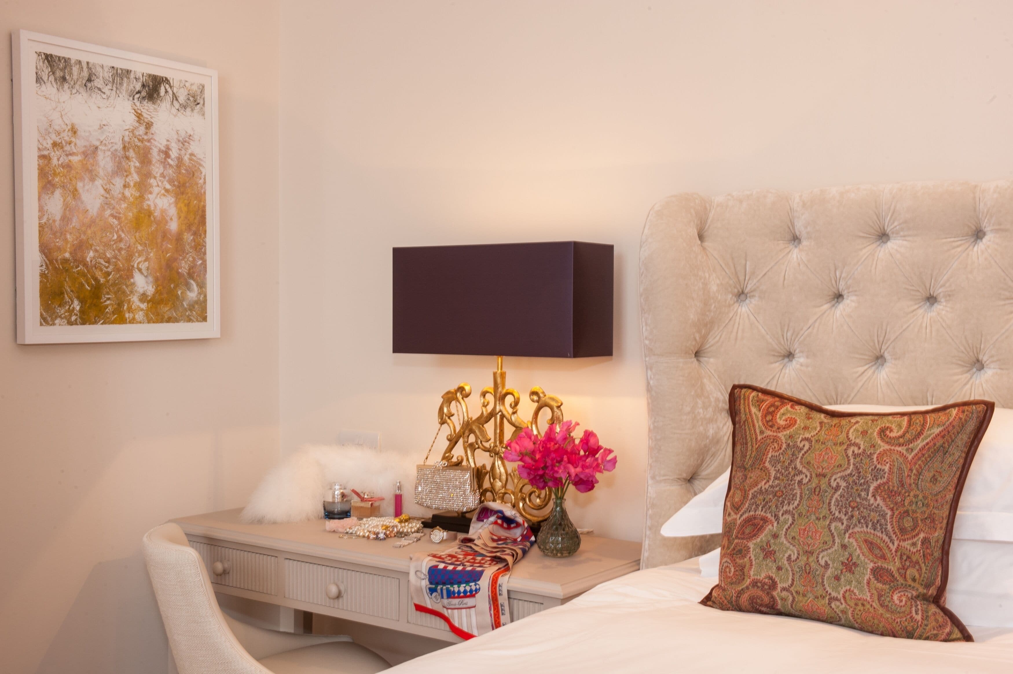 A neatly arranged bedroom with a tufted headboard, a bedside table with a lamp, a framed artwork, and a decorative pillow.
