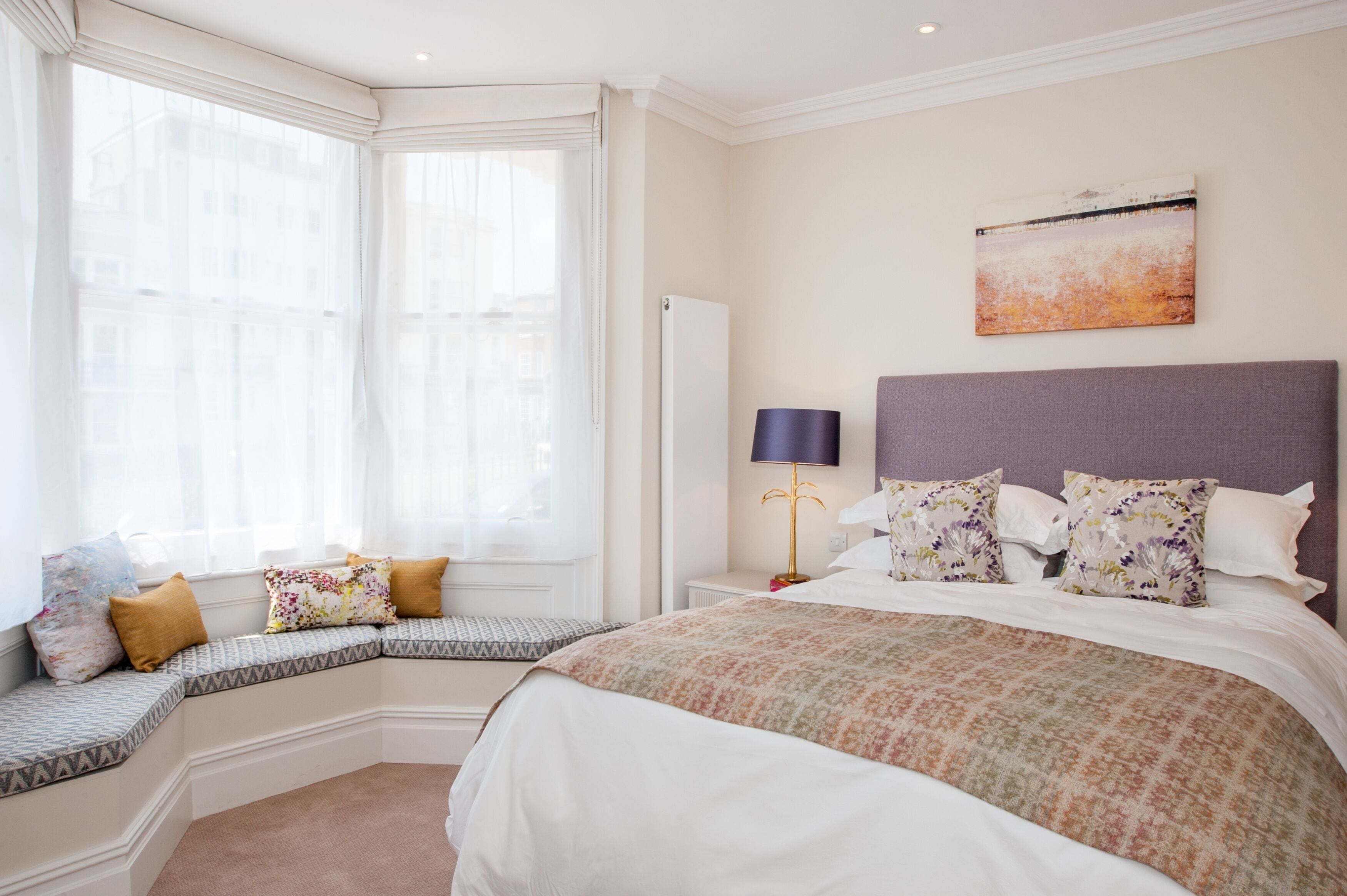 A bedroom with a large bed, a nightstand, a lamp, and a window seat with cushions.