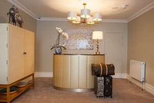A reception area with a curved reception desk, a chandelier, a large abstract painting, a lamp, and a luggage stand with a suitcase and a Louis Vuitton bag.