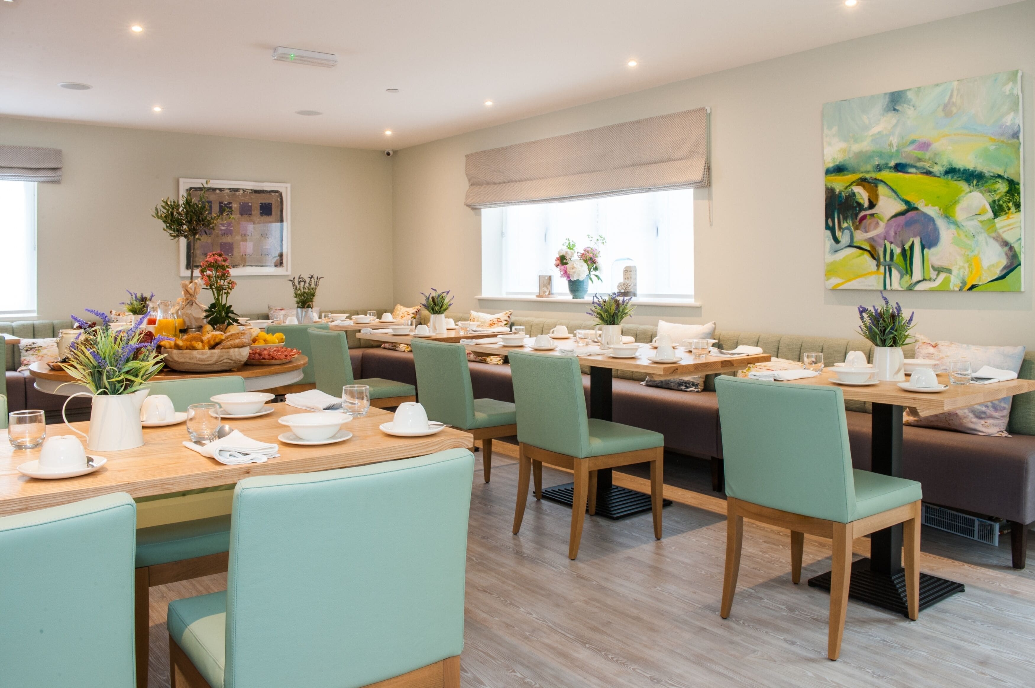 Un espace repas avec plusieurs tables et chaises, un grand tableau accroché au mur et une fenêtre donnant sur l’extérieur.