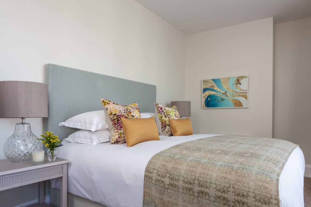 Een netjes opgemaakt slaapkamer met een bed, kussens, een nachtkastje met een lamp en een schilderij aan de muur.