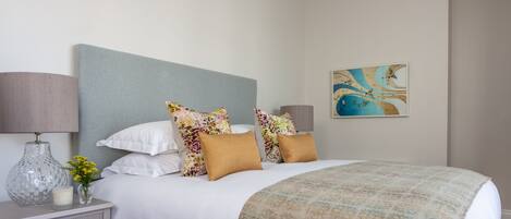 Un dormitorio ordenado con cama, almohadas, una mesita de noche con lámpara y un cuadro en la pared.