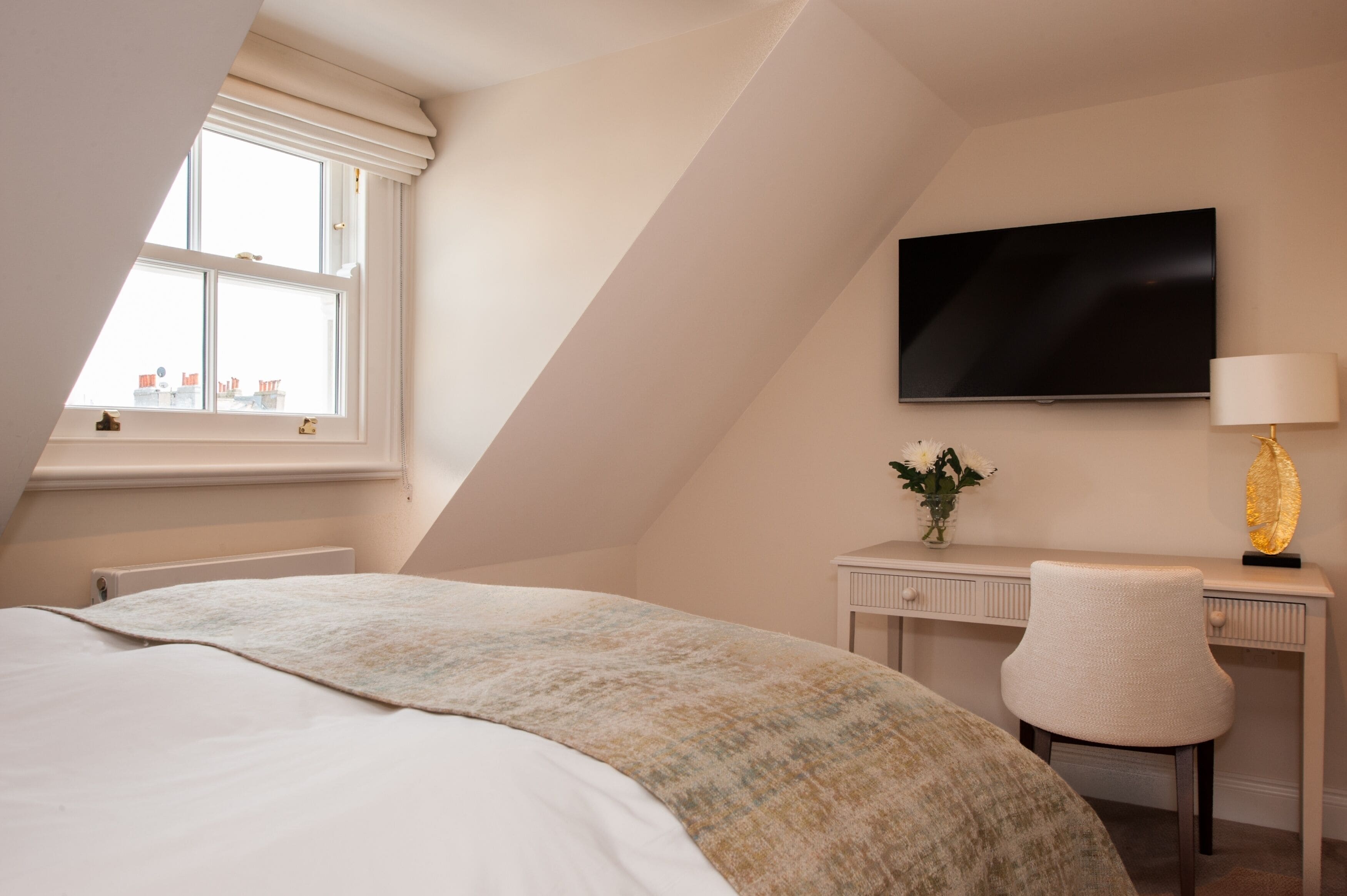 A bedroom with a large bed, a desk with a chair, a flat-screen TV, and a window with a view of buildings.