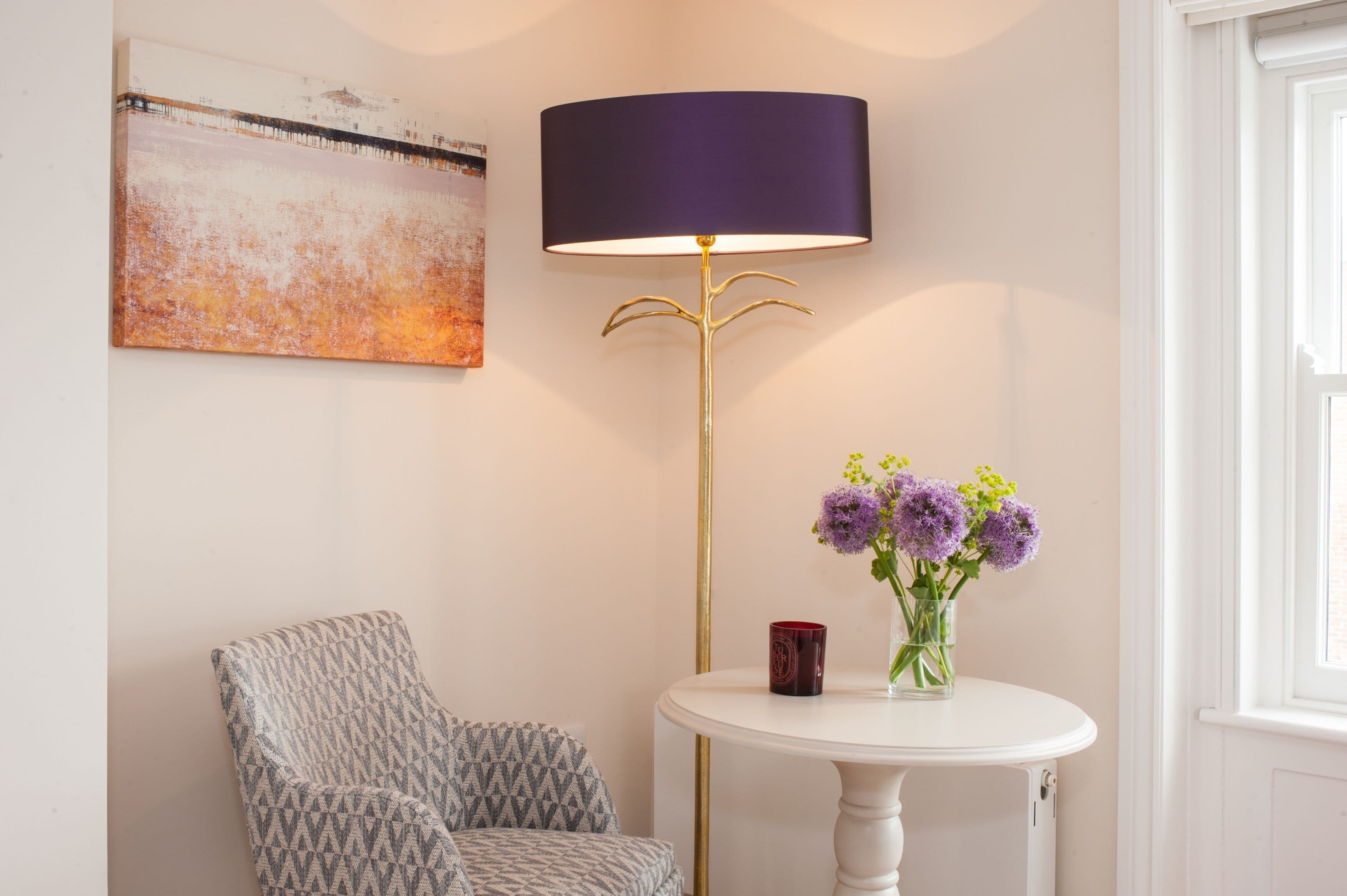 Una habitación con un sillón estampado, una mesa redonda con un jarrón de flores, una lámpara de pie y un cuadro en la pared.