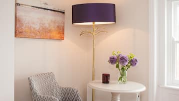 Una habitación con un sillón estampado, una mesa redonda con un jarrón de flores, una lámpara de pie y un cuadro en la pared.