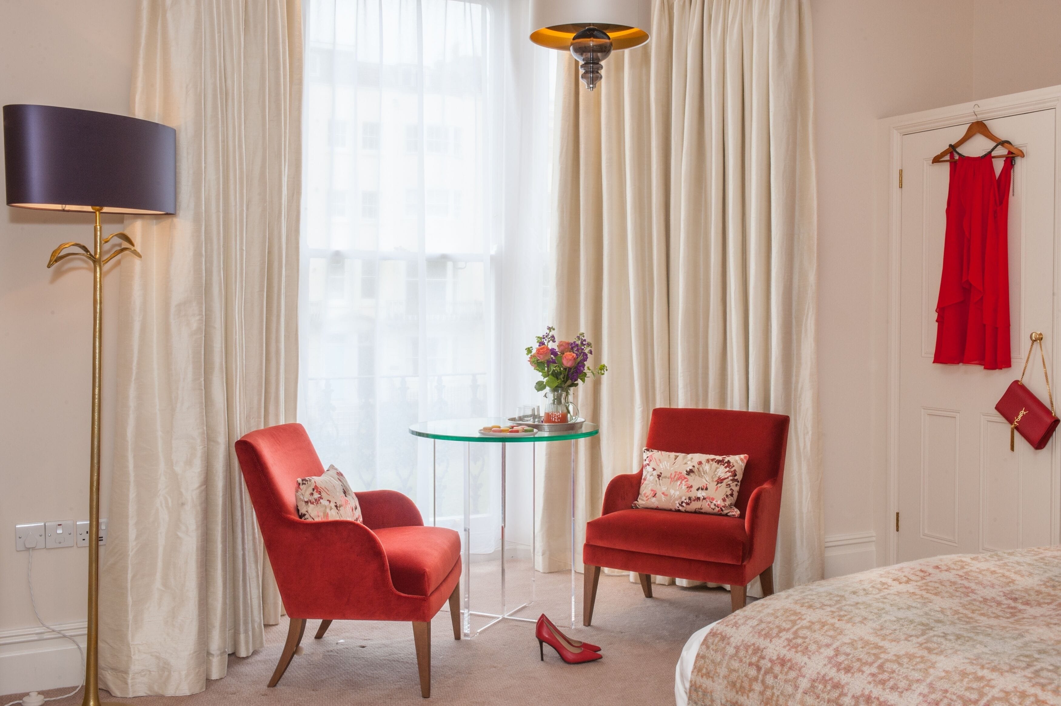 A hotel room with two red armchairs, a small round table with a vase of flowers, a lamp, and a bed with a floral bedspread.