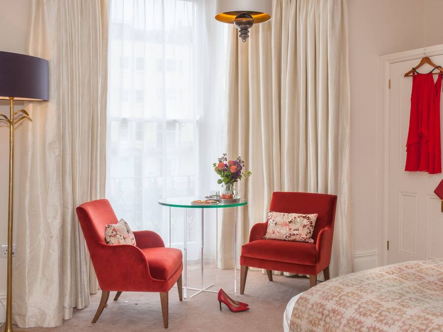 A hotel room with two red armchairs, a small round table with a vase of flowers, a lamp, and a bed with a floral bedspread.