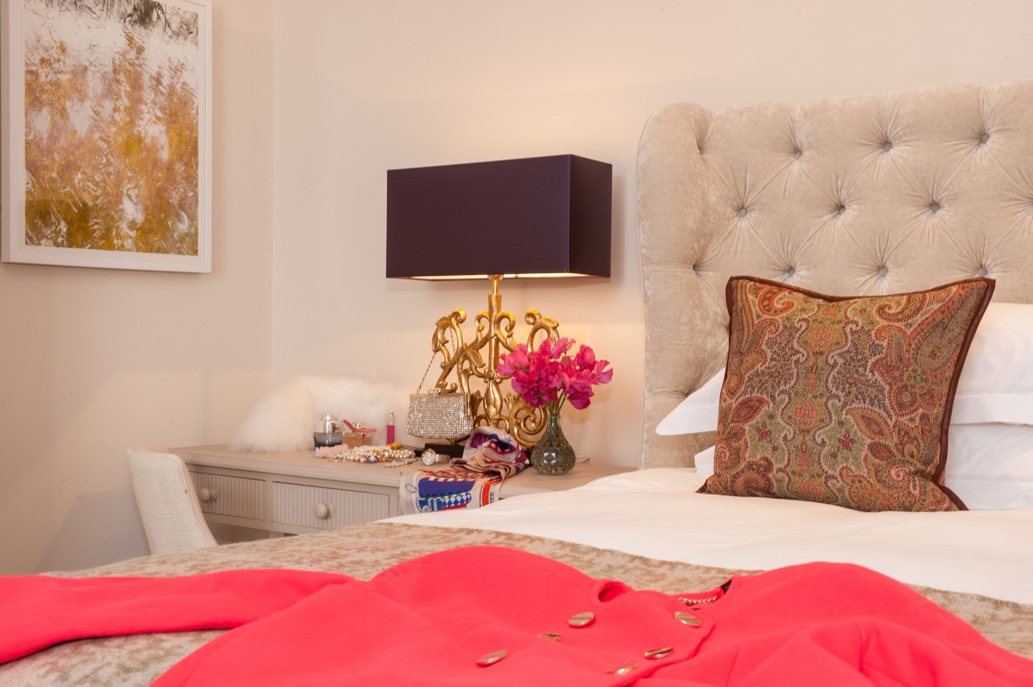 A neatly arranged bedroom with a tufted headboard, a bedside table with a lamp, a decorative vase, and a plush red blanket.