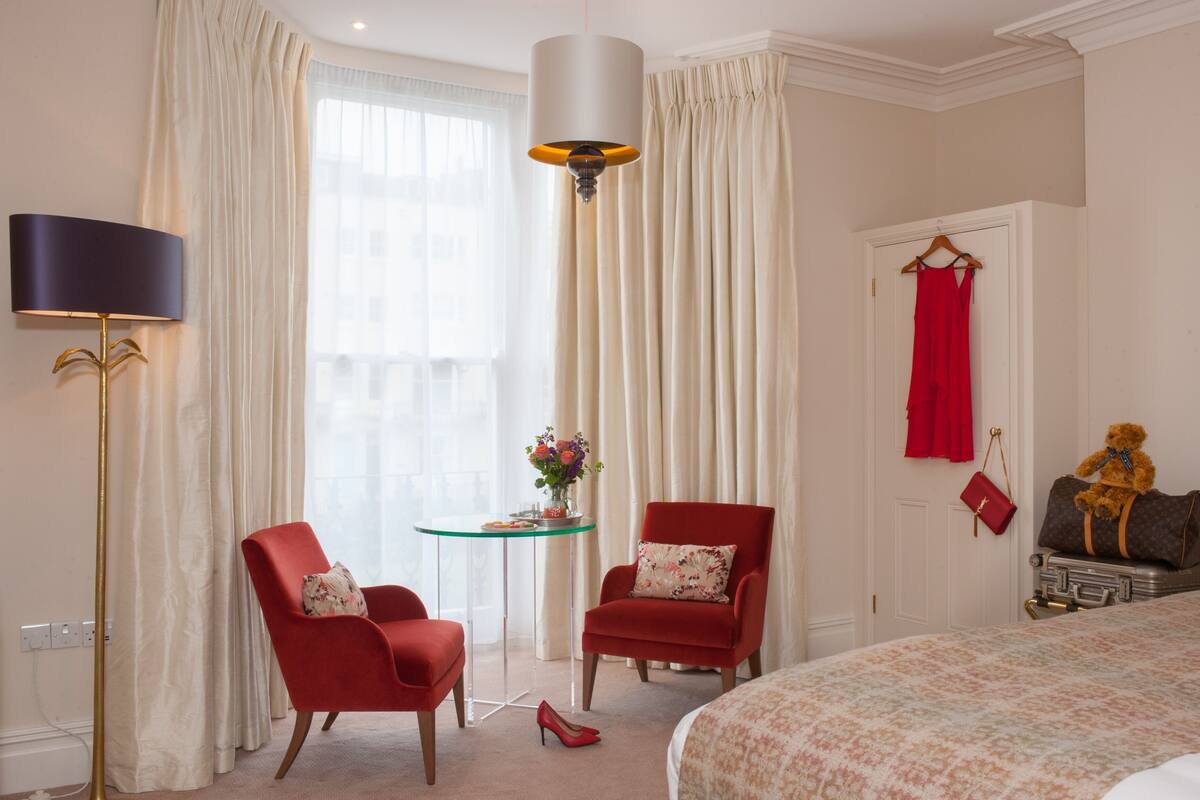 Een hotelkamer met twee rode fauteuils, een glazen tafel met een bloevas, een bed met een bloemenprintdeken, een rode jurk aan een kapstok en een teddybeer op een koffer.
