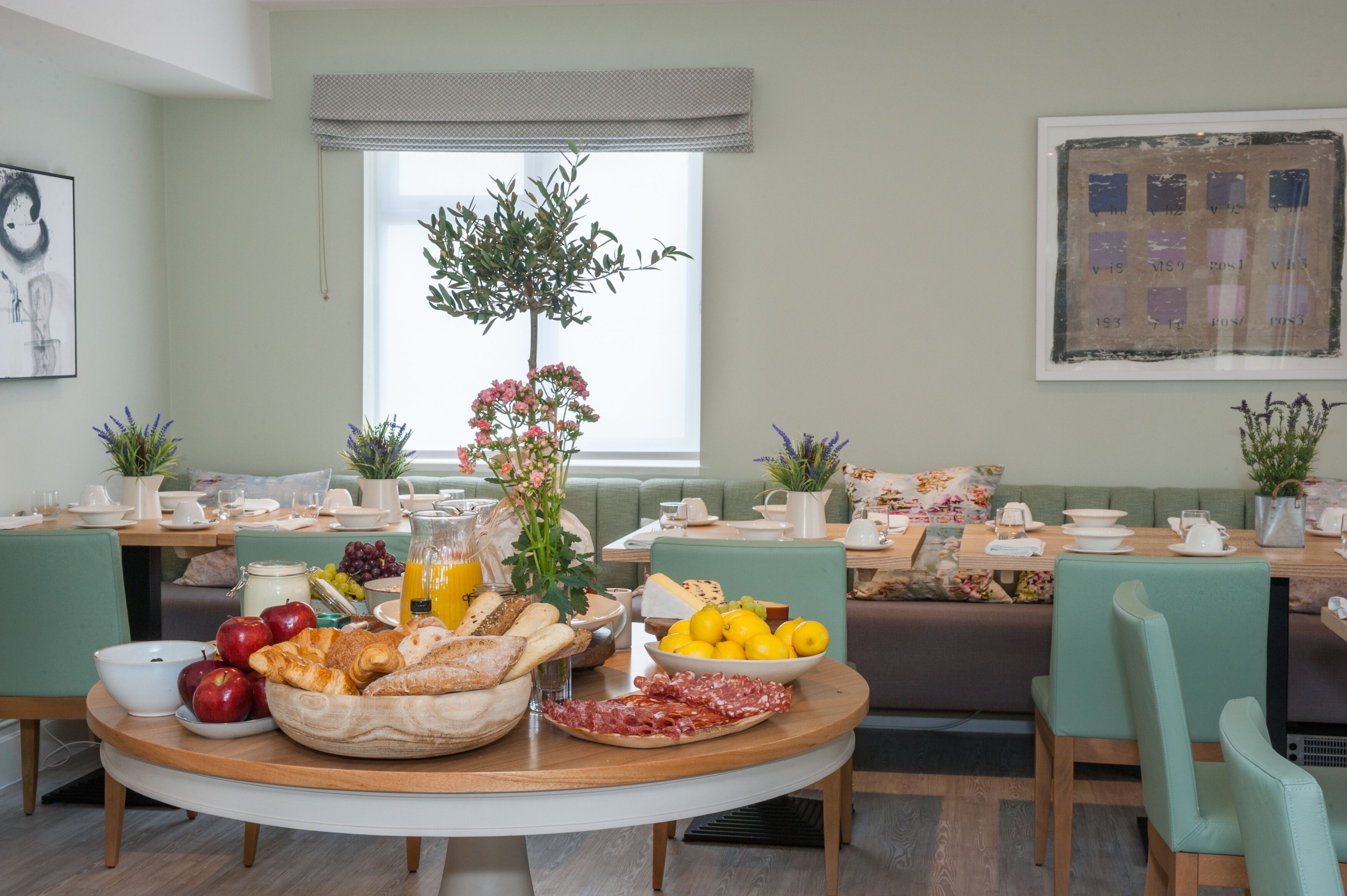 A breakfast setup with a variety of foods and drinks on a round table, a green sofa, and a window with blinds.