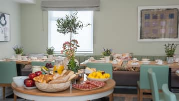 A breakfast setup with a variety of foods and drinks on a round table, a green sofa, and a window with blinds.