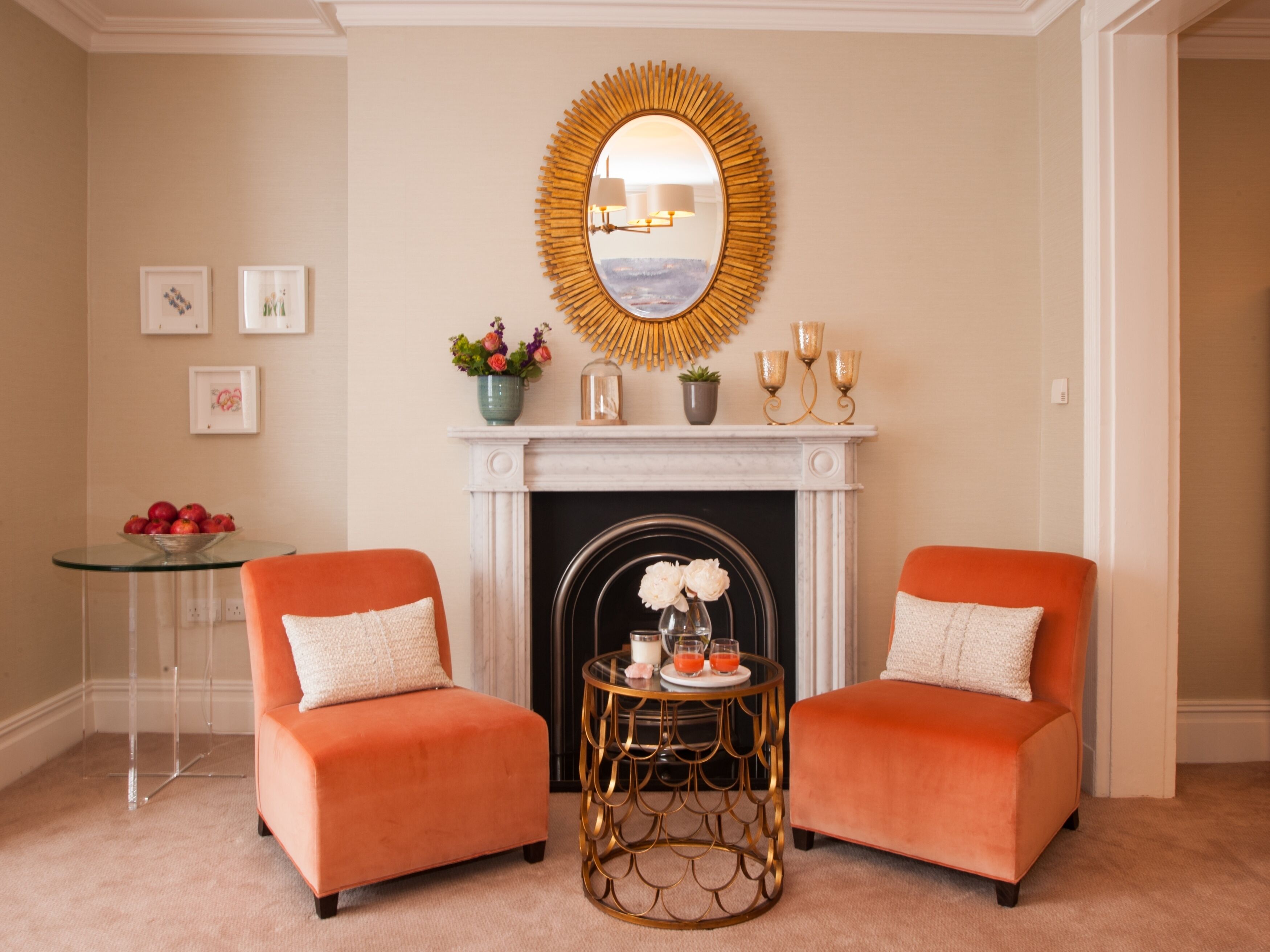 A room with two orange armchairs, a round mirror above a fireplace, and a small table with a vase and candles.