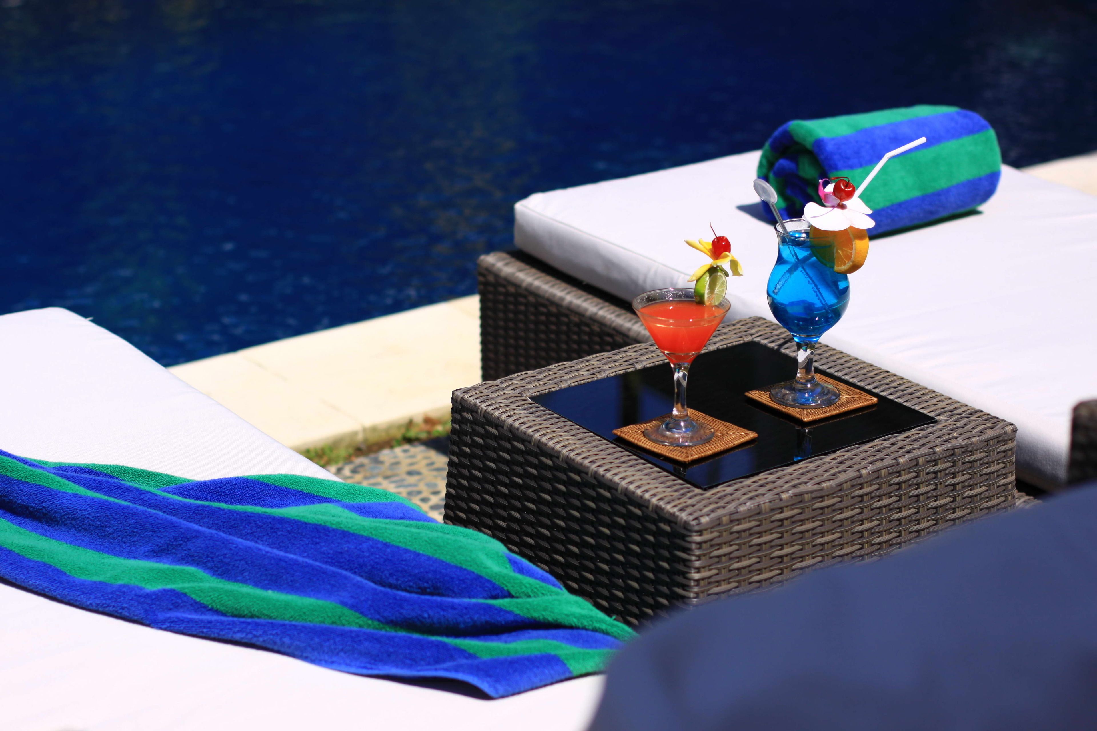 A poolside area with lounge chairs, a wicker table with drinks, and a blue and green towel.
