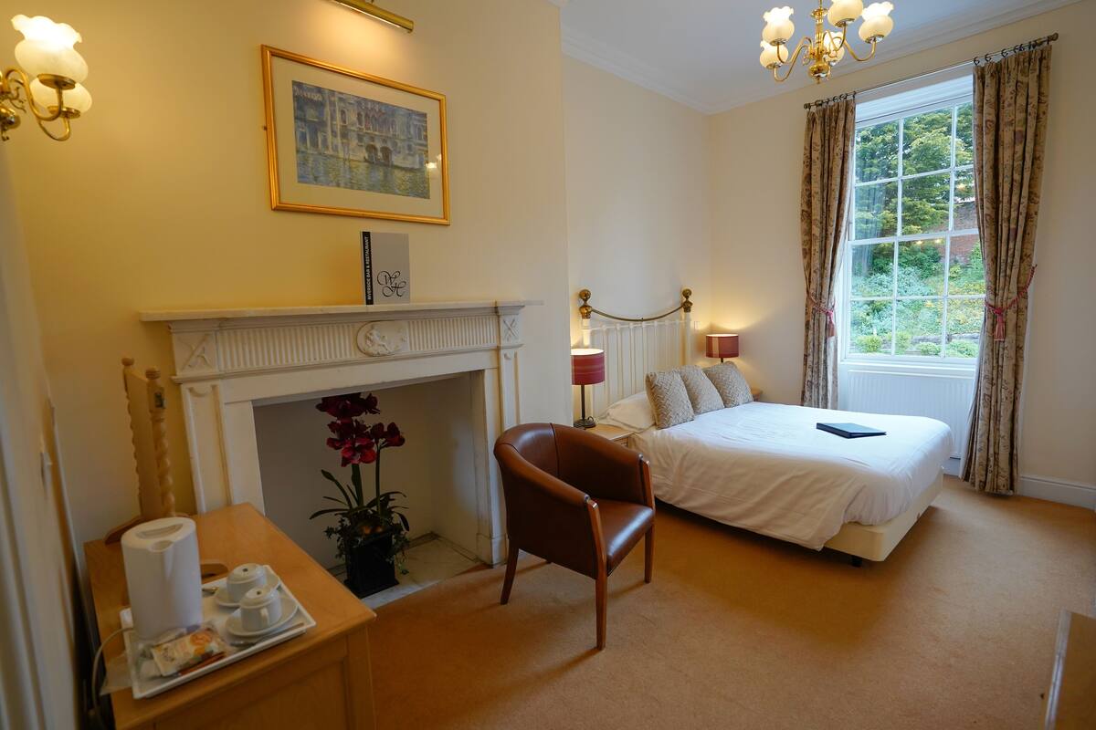 A bedroom with a bed, a chair, a bedside table with a tray, a fireplace, a window with curtains, and a chandelier.