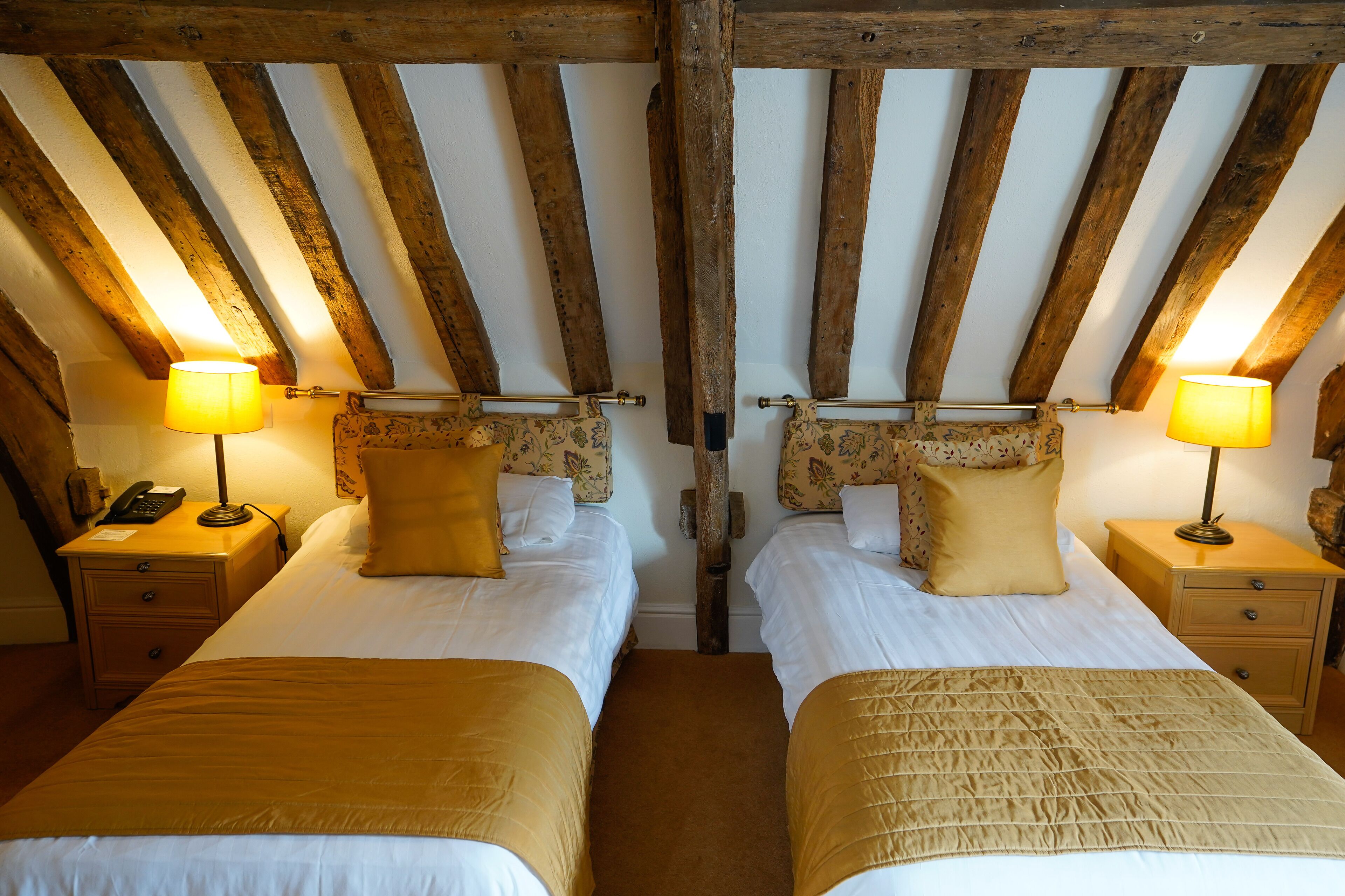 A room with two beds, wooden beams on the ceiling, bedside lamps, and a telephone on the nightstand.