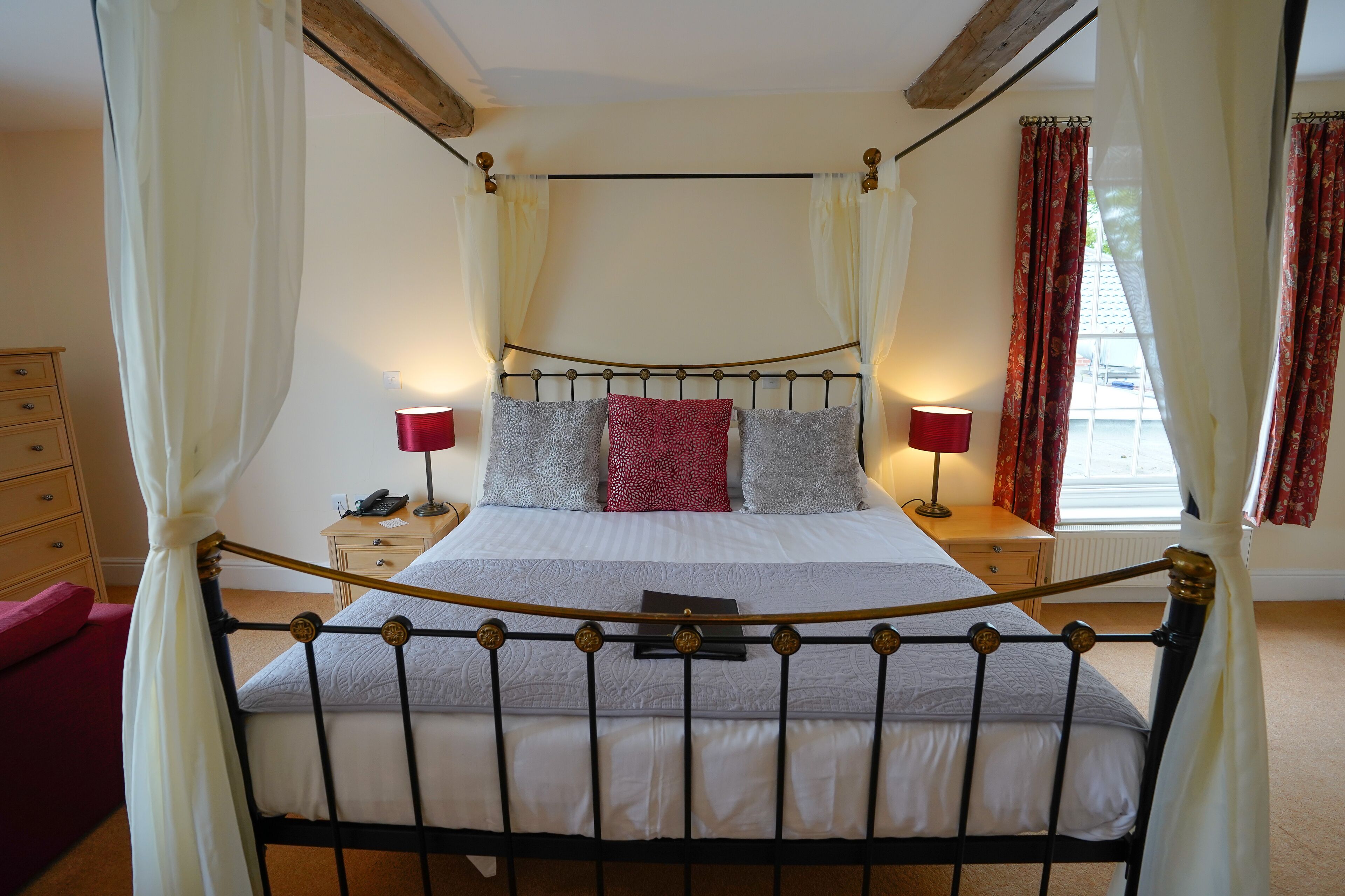 Een bed met hemelbed met een metalen frame, wit beddengoed en rode en grijze kussens. Aan een kant staan nachtkastjes met lampen en een telefoon. De kamer heeft een houten plafond en een raam met rode gordijnen.