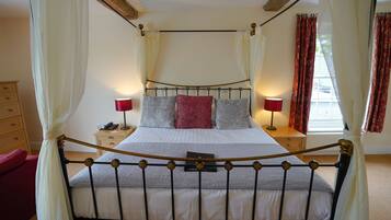 Een bed met hemelbed met een metalen frame, wit beddengoed en rode en grijze kussens. Aan een kant staan nachtkastjes met lampen en een telefoon. De kamer heeft een houten plafond en een raam met rode gordijnen.