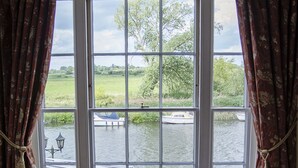 A room with a large window overlooking a river, a wooden table with a book, and curtains. - Waveney House Hotel (Beccles)