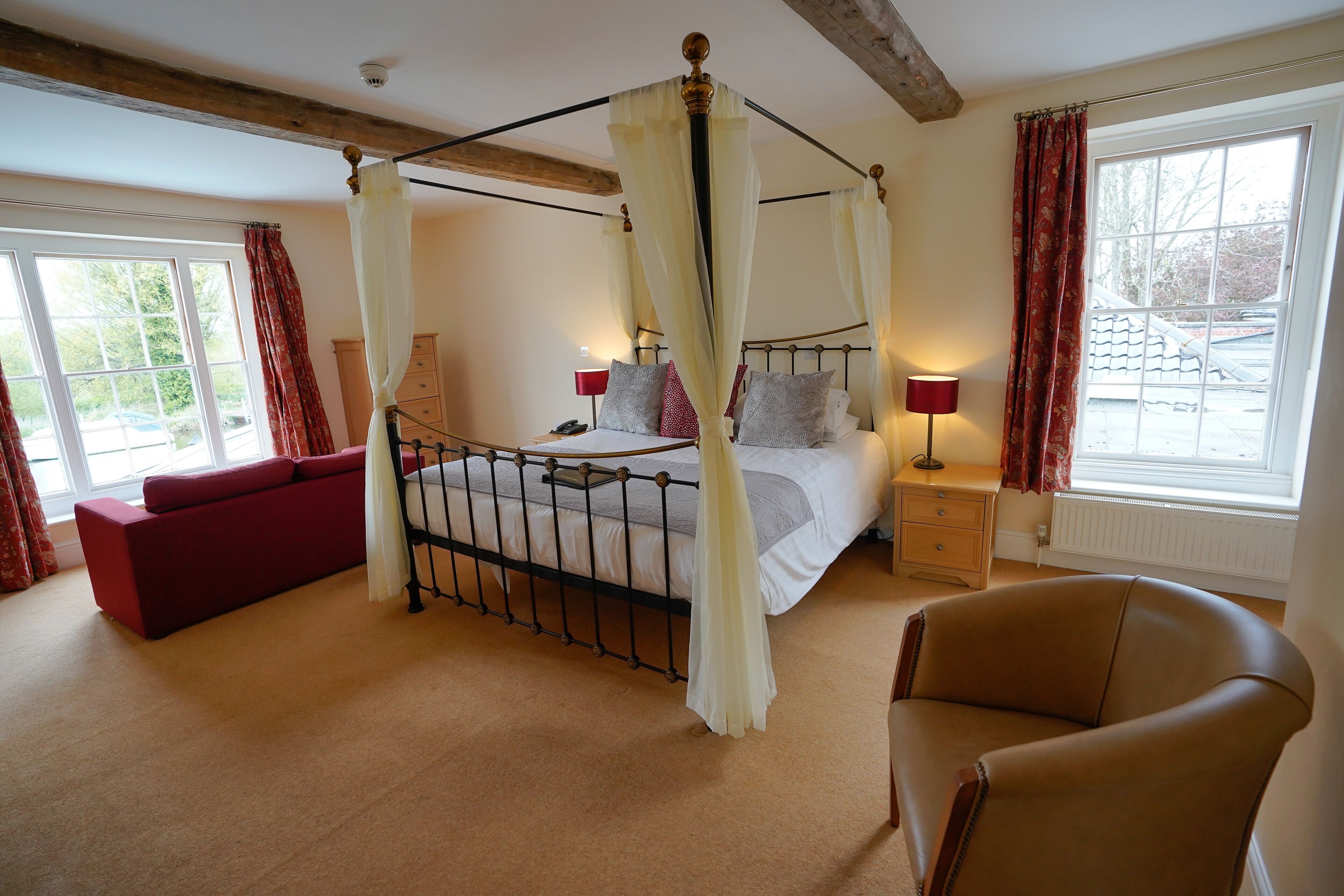 A bedroom with a four-poster bed, a red sofa, a brown armchair, and a window with curtains.