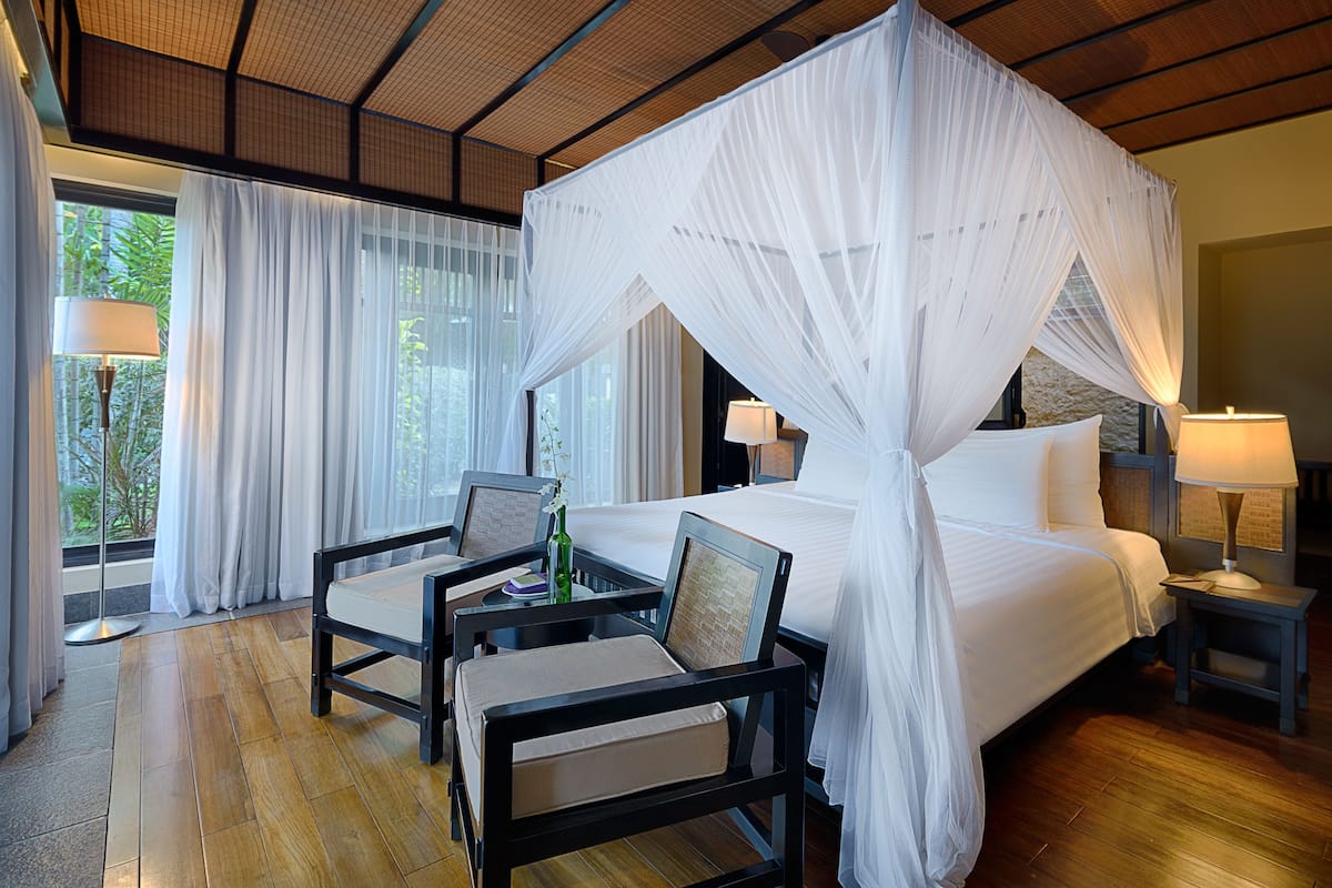 A room with a four-poster bed, a sitting area with chairs, a small table, and a nightstand with a lamp.