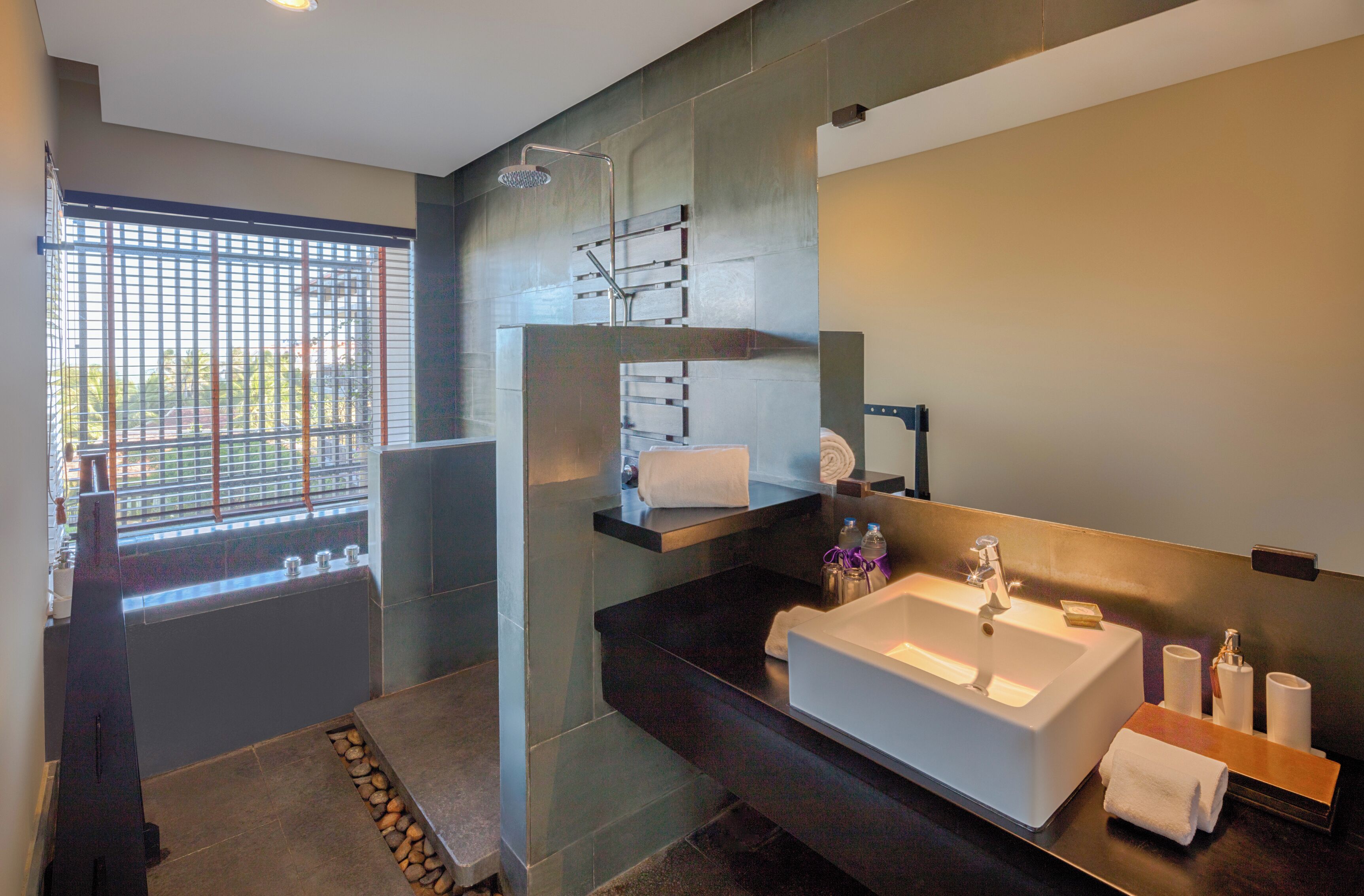 A modern bathroom with a large sink, a shower, and a window with blinds.