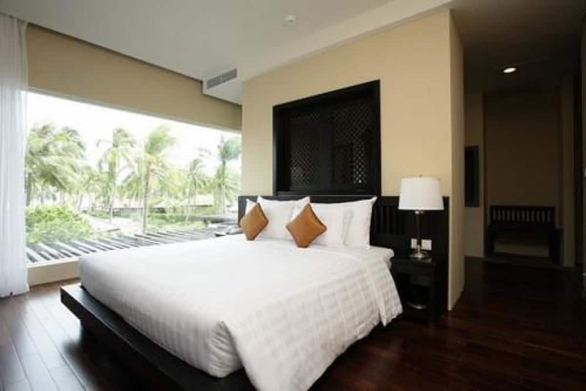 A hotel room with a large bed, a nightstand with a lamp, a view of palm trees, and a dark wooden floor.