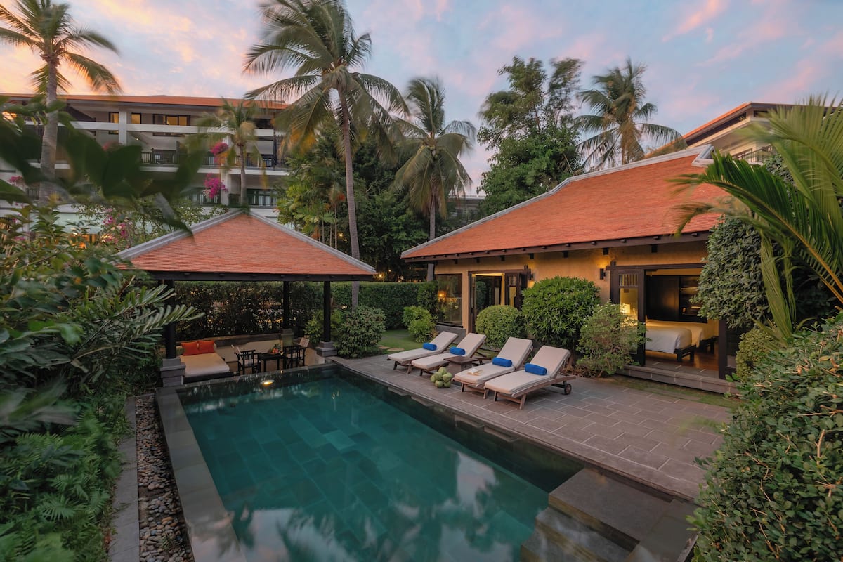 A pool area with lounge chairs and umbrellas, surrounded by palm trees and a building with a red roof.