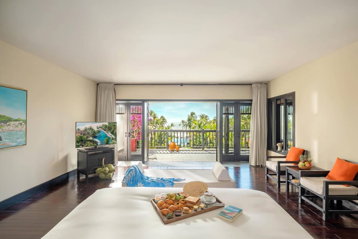 A hotel room with a bed, a dining table with breakfast, a TV, and a balcony with a view of palm trees and the sea.