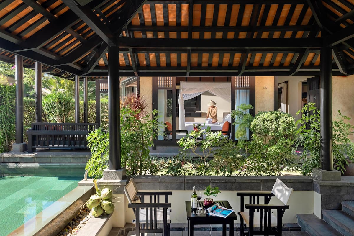 A covered outdoor seating area with a pool, wooden furniture, and lush greenery.