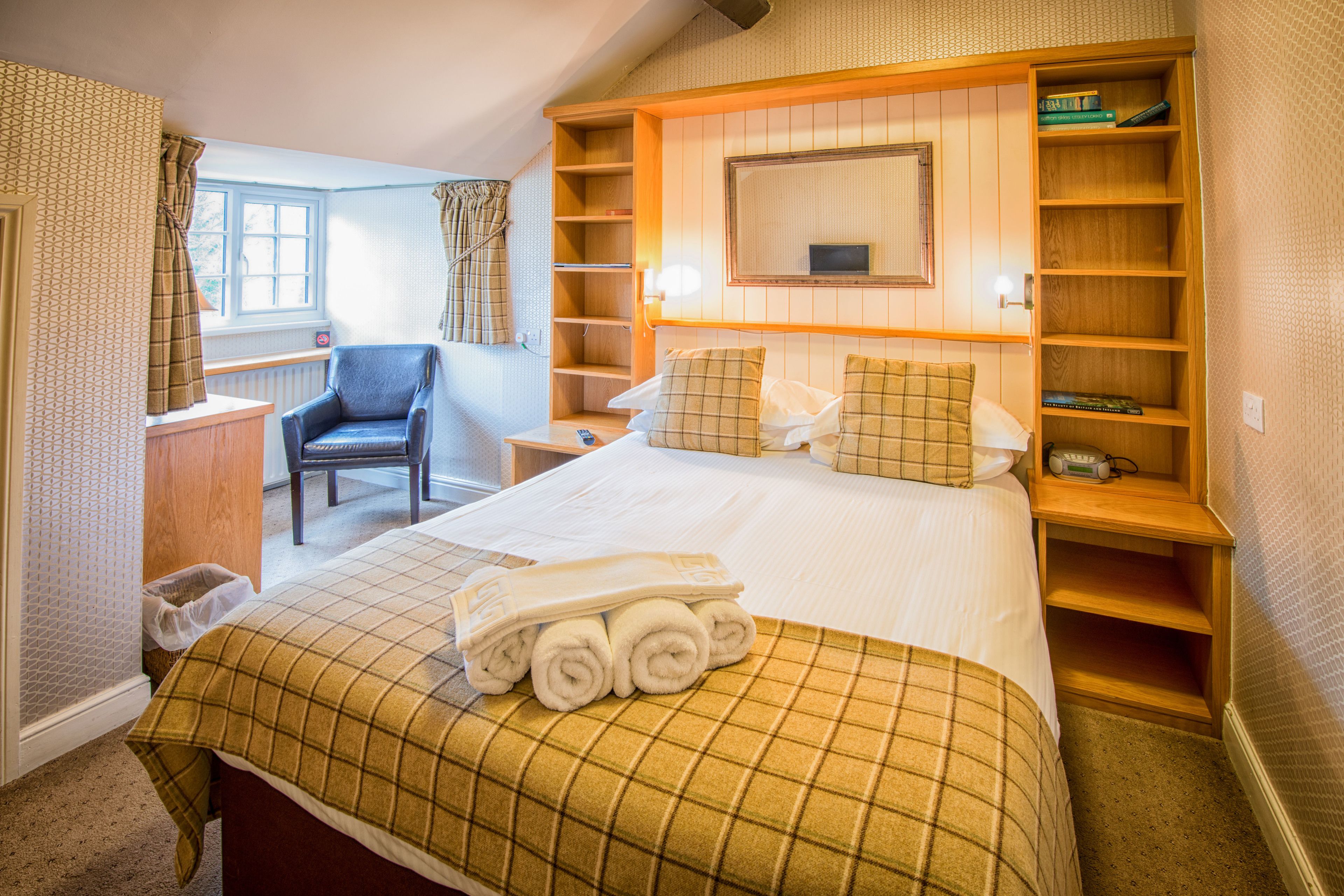 A hotel room with a bed, a chair, a bedside table, a bookshelf, and a window with curtains.