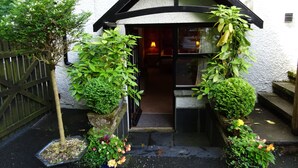 A quaint building with a black and white exterior, a wooden door, and a small porch area with potted plants and flowers. - Woodlands Guest House Windermere (Windermere)