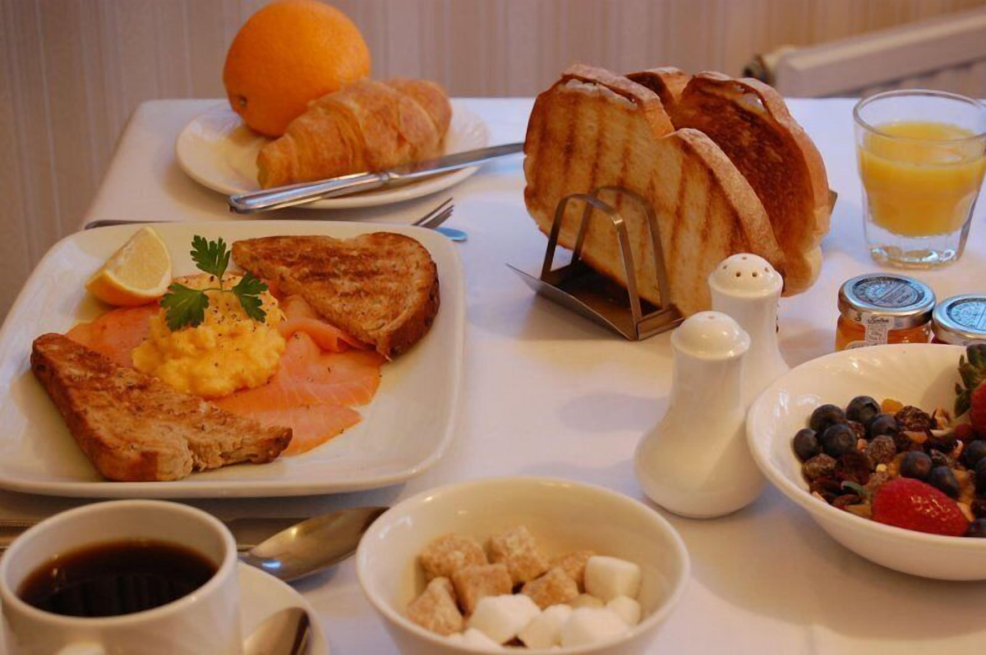 A breakfast table with toast, scrambled eggs, smoked salmon, croissants, a glass of orange juice, a bowl of fruit, and condiments.