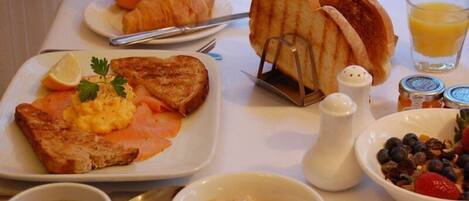 A breakfast table with toast, scrambled eggs, smoked salmon, croissants, a glass of orange juice, a bowl of fruit, and condiments.