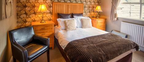 Un dormitorio con cabecera de madera, cama con manta marrón, dos lámparas de noche, una mesita de noche de madera, una silla y papel tapiz floral.