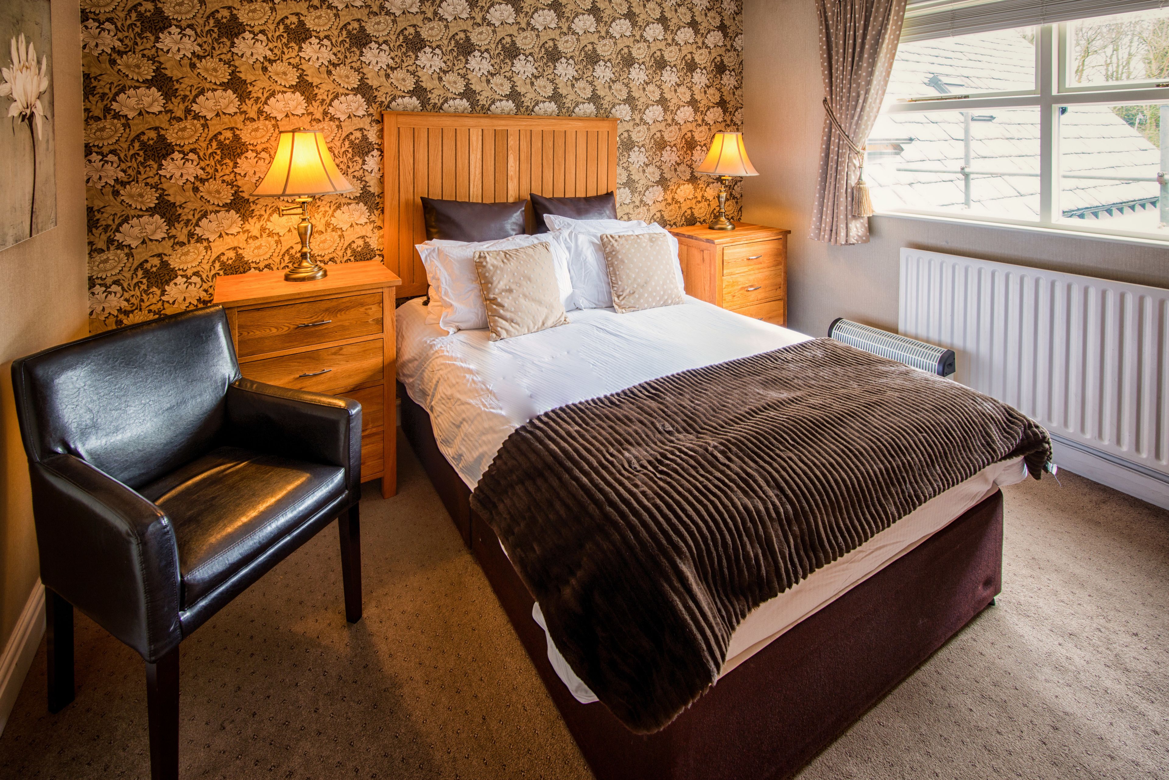 A bedroom with a bed, two bedside tables, a chair, a radiator, and a window with curtains.
