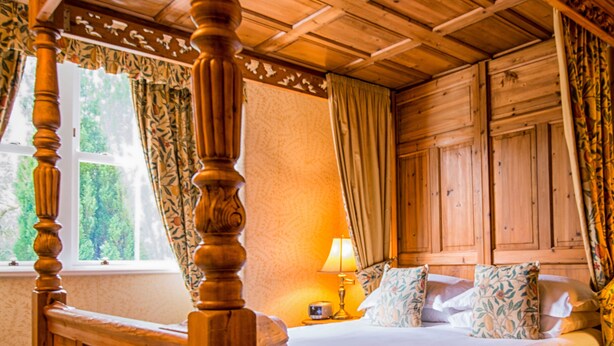 A four-poster bed with a wooden headboard and carved details, a bedside table with a lamp, and a window with curtains. - Woodlands Guest House Windermere (Windermere)