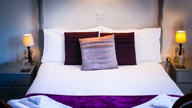 A neatly made bed with white and purple bedding, two bedside lamps, and a nightstand with a glass on it. - The Hostelrie at Goodrich (Ross-on-Wye)